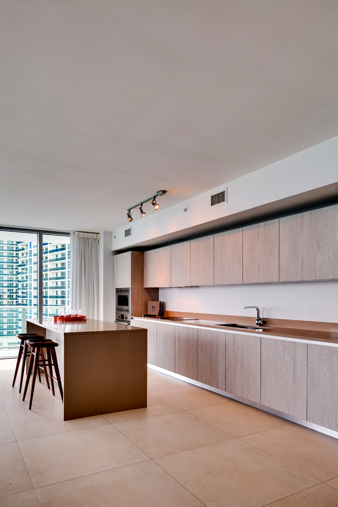 This is an example of a large modern single-wall kitchen/diner in Miami with a submerged sink, flat-panel cabinets, light wood cabinets, white splashback, stainless steel appliances, an island, beige floors and beige worktops.