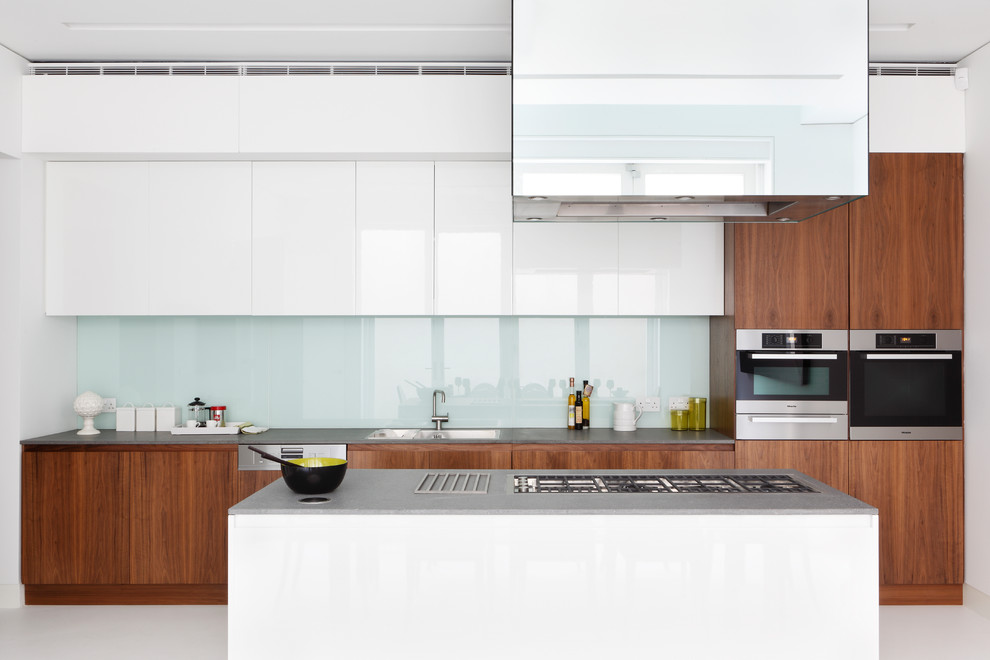 Photo of a contemporary galley kitchen in Other with a built-in sink, flat-panel cabinets, medium wood cabinets, glass sheet splashback, an island, white floors and grey worktops.