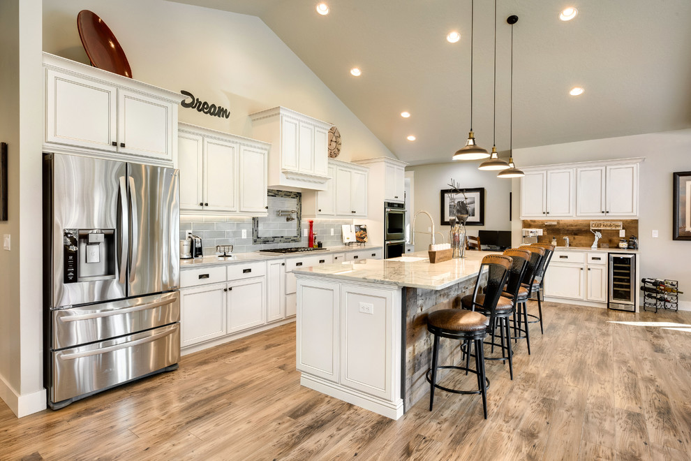 Example of a large farmhouse l-shaped medium tone wood floor and brown floor open concept kitchen design in Orange County with an undermount sink, shaker cabinets, white cabinets, gray backsplash, subway tile backsplash, stainless steel appliances, an island and white countertops