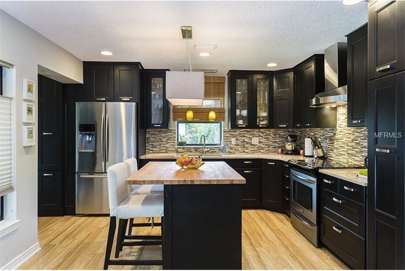 This is an example of a medium sized bohemian l-shaped kitchen/diner in Orlando with a double-bowl sink, recessed-panel cabinets, black cabinets, composite countertops, multi-coloured splashback, glass tiled splashback, stainless steel appliances, light hardwood flooring and an island.