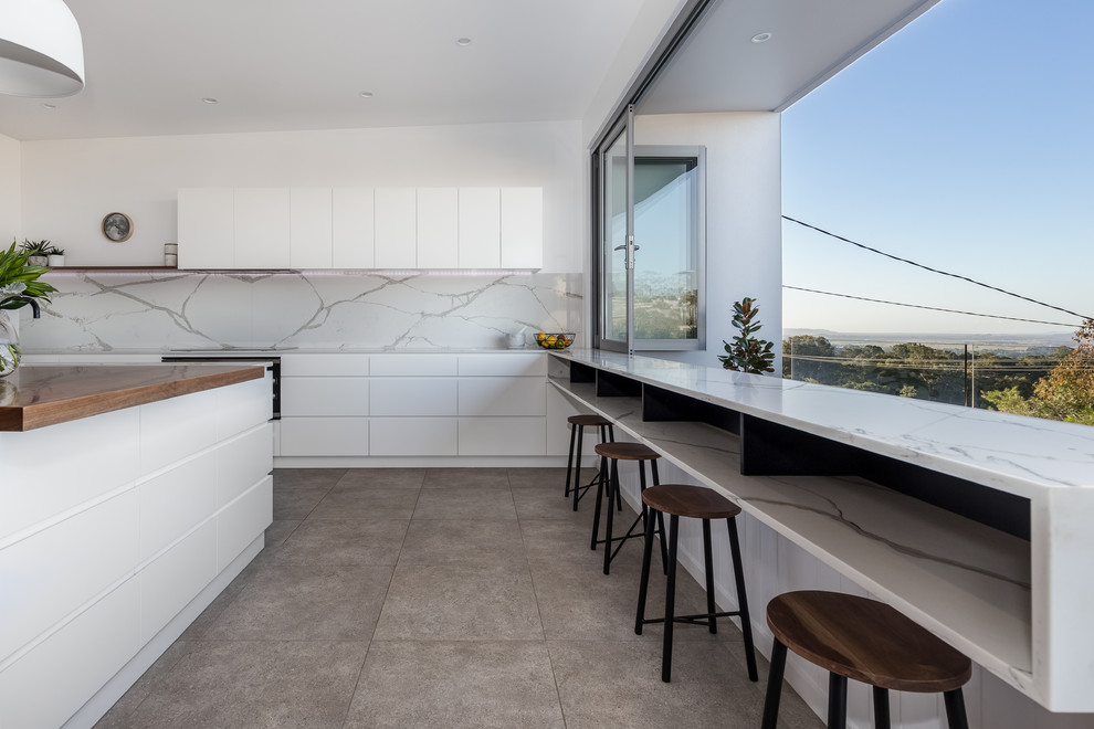 Modern inredning av ett stort vit vitt kök, med en köksö, en enkel diskho, släta luckor, vita skåp, bänkskiva i kalksten, grått stänkskydd, stänkskydd i marmor, rostfria vitvaror och beiget golv