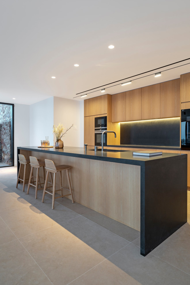 This is an example of an expansive contemporary galley open plan kitchen with granite worktops, black splashback, stainless steel appliances, marble flooring, an island, beige floors, black worktops, a submerged sink, flat-panel cabinets and medium wood cabinets.