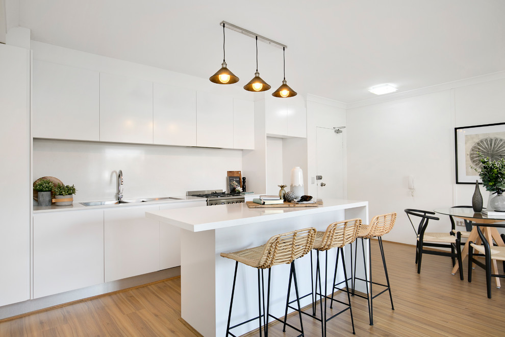 Inspiration for a contemporary single-wall kitchen/diner in Sydney with a double-bowl sink, flat-panel cabinets, white cabinets, composite countertops, white splashback, stainless steel appliances, laminate floors, an island, brown floors and white worktops.