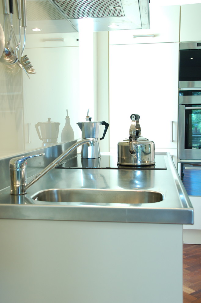 Foto på ett nordiskt kök, med en integrerad diskho, släta luckor, vita skåp, bänkskiva i rostfritt stål, glaspanel som stänkskydd, rostfria vitvaror och mörkt trägolv
