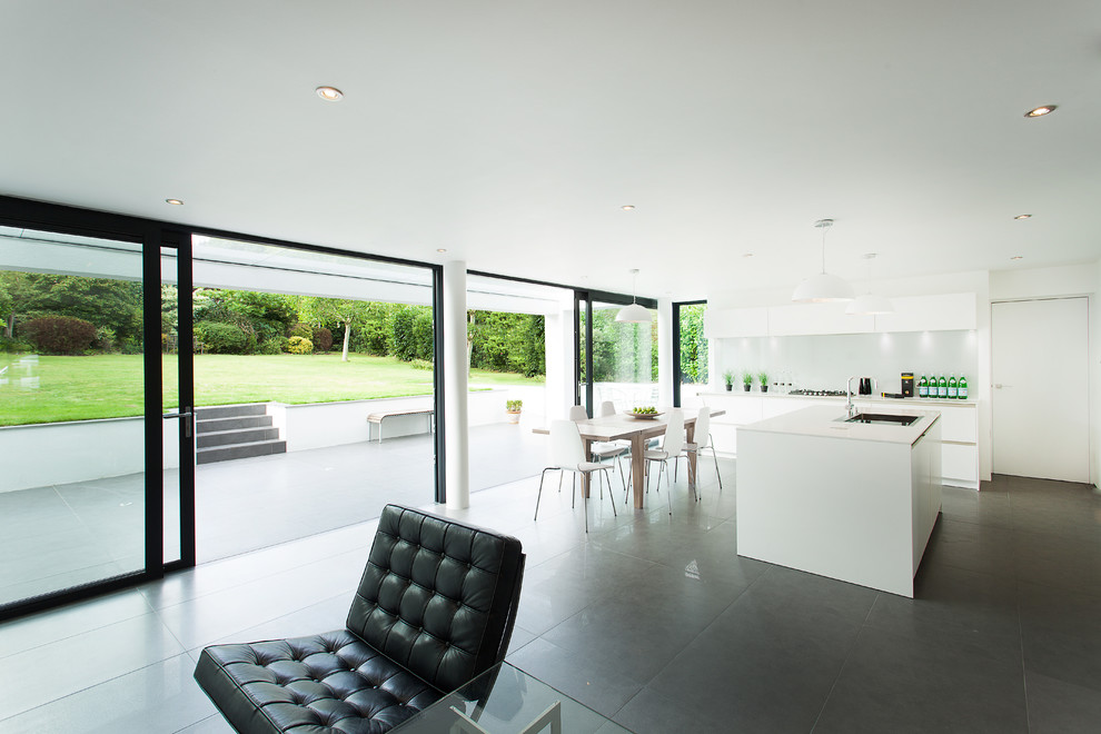 Large modern single-wall open plan kitchen in Hampshire with a submerged sink, flat-panel cabinets, white cabinets, composite countertops, grey splashback, glass sheet splashback, stainless steel appliances and an island.