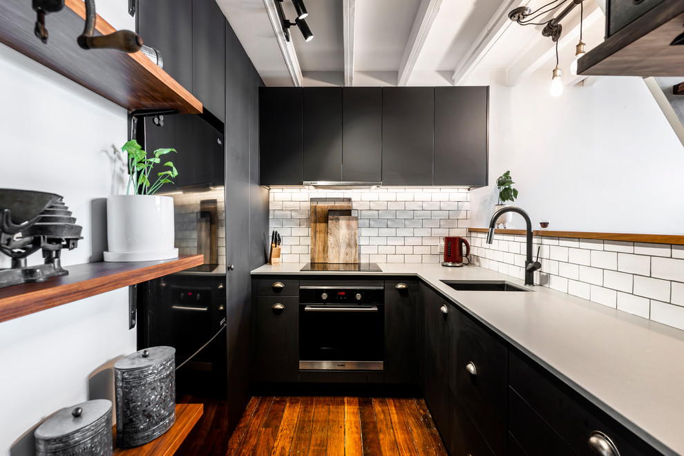 Example of an urban u-shaped dark wood floor and brown floor kitchen design in Brisbane with an undermount sink, flat-panel cabinets, black cabinets, white backsplash, subway tile backsplash, black appliances, a peninsula and gray countertops