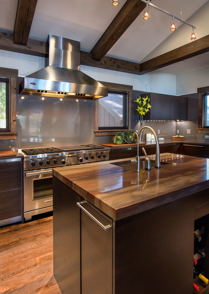 Foto på ett stort industriellt kök, med en undermonterad diskho, släta luckor, skåp i mörkt trä, grått stänkskydd, glaspanel som stänkskydd, rostfria vitvaror, mellanmörkt trägolv och en köksö