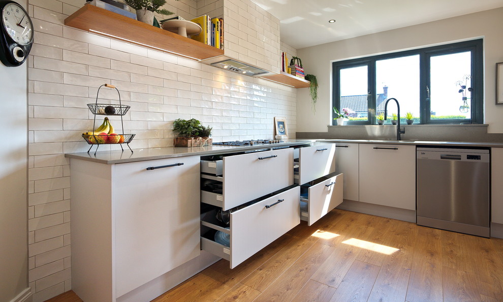 Medium sized industrial l-shaped kitchen/diner in Other with a built-in sink, flat-panel cabinets, grey cabinets, quartz worktops, white splashback, ceramic splashback, stainless steel appliances, laminate floors, an island, brown floors and grey worktops.