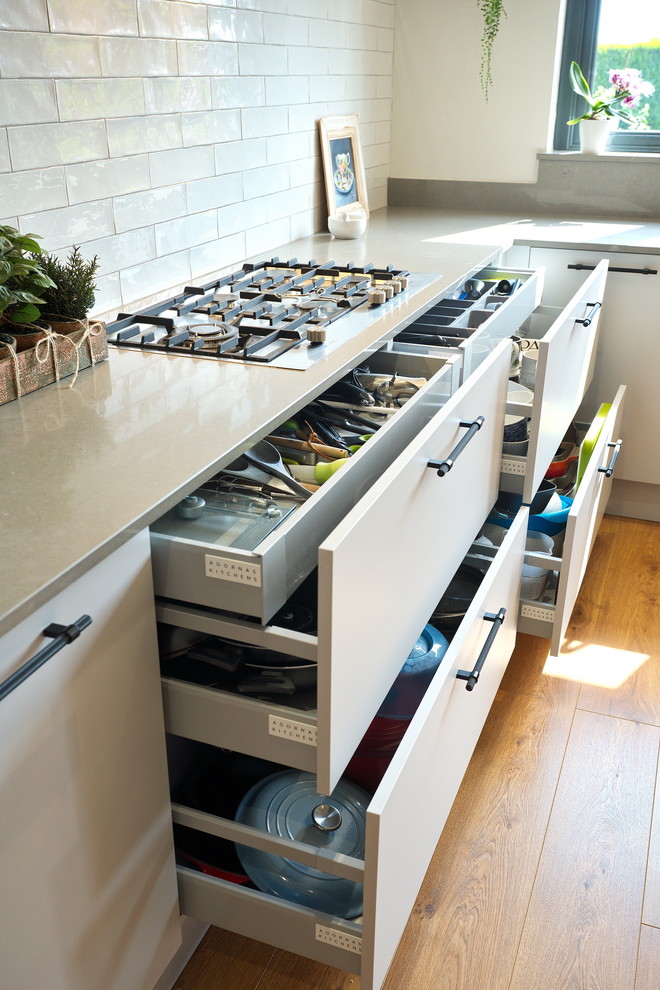 This is an example of a medium sized urban l-shaped kitchen/diner in Other with a built-in sink, flat-panel cabinets, grey cabinets, quartz worktops, white splashback, ceramic splashback, stainless steel appliances, laminate floors, an island, brown floors and grey worktops.