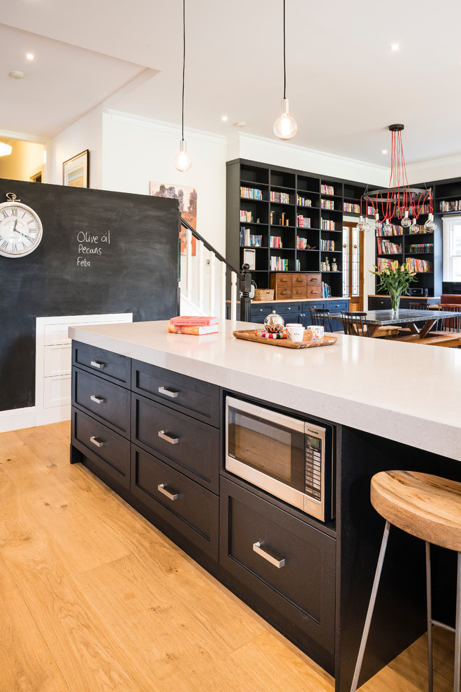 Large modern l-shaped open plan kitchen in Melbourne with recessed-panel cabinets, black cabinets, engineered stone countertops, white splashback, an island, a double-bowl sink, metro tiled splashback, stainless steel appliances and light hardwood flooring.