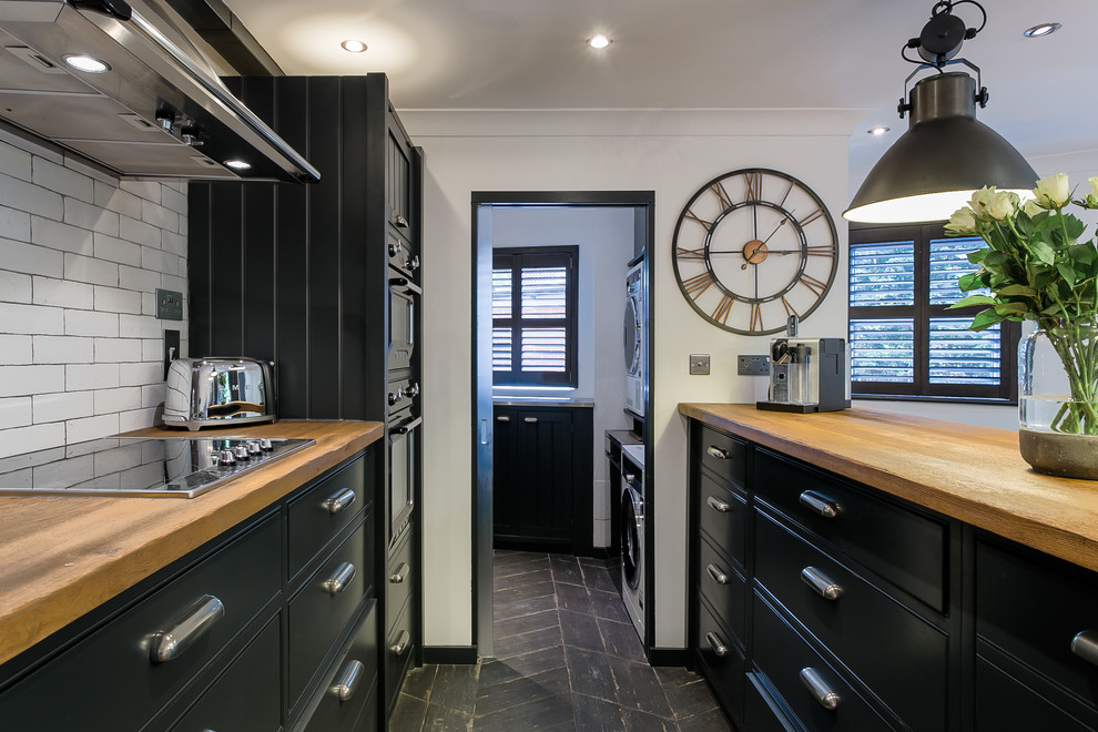 Immagine di una cucina industriale con ante lisce, ante nere, top in legno, paraspruzzi bianco, elettrodomestici da incasso, pavimento nero e top marrone