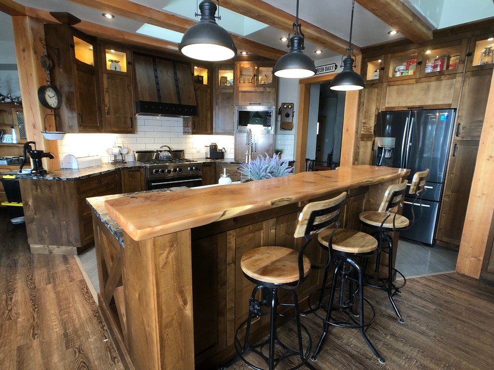 This is an example of a large industrial l-shaped kitchen/diner in Vancouver with a belfast sink, shaker cabinets, medium wood cabinets, white splashback, metro tiled splashback, stainless steel appliances, light hardwood flooring, an island, brown floors and black worktops.