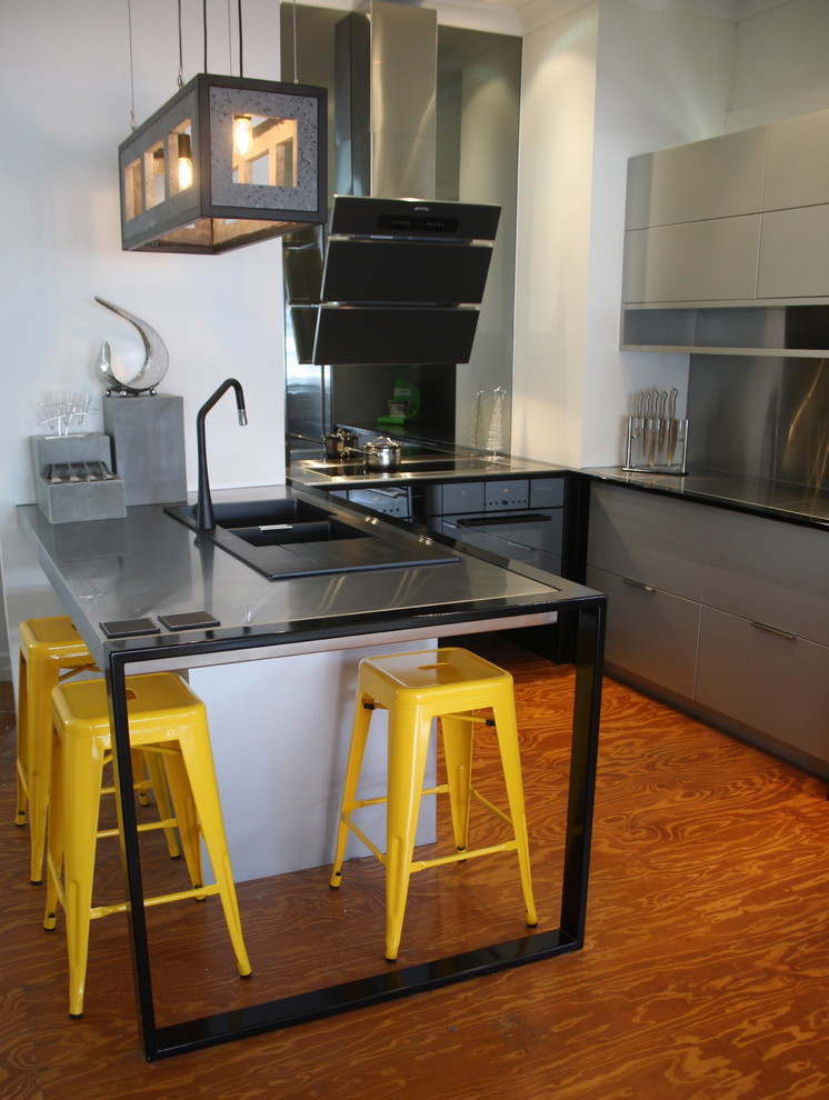 Bild på ett avskilt, litet industriellt u-kök, med en dubbel diskho, släta luckor, grå skåp, bänkskiva i rostfritt stål, grått stänkskydd, stänkskydd i metallkakel, svarta vitvaror, mellanmörkt trägolv och en köksö