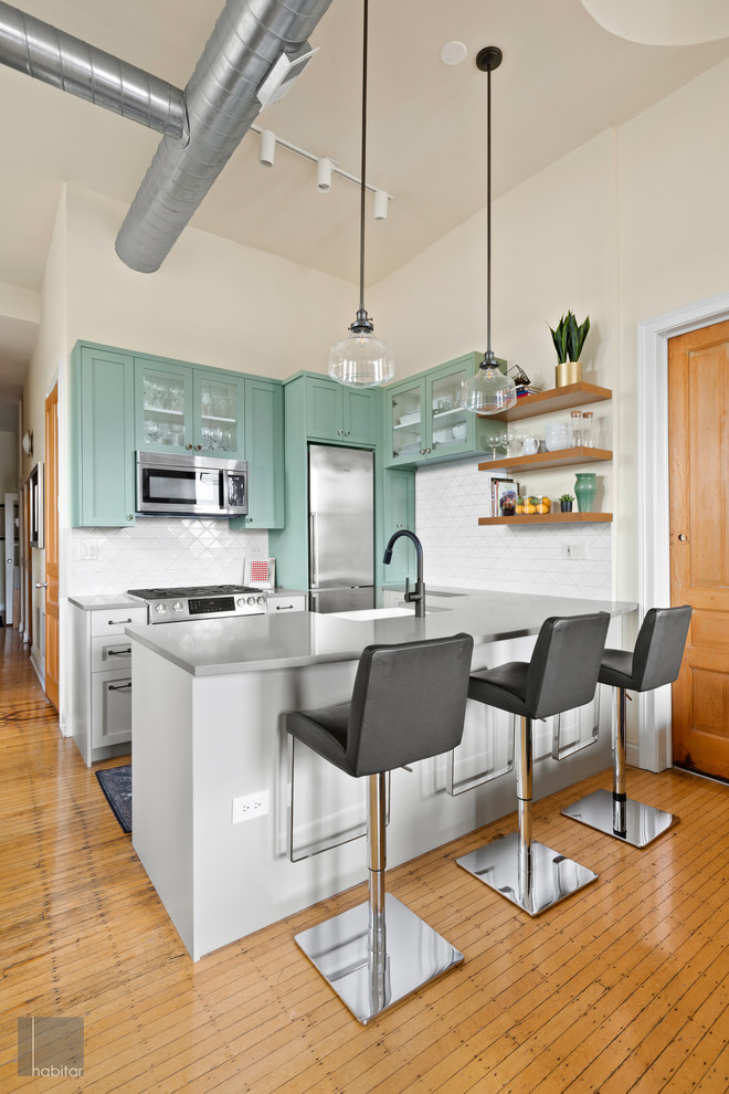 Medium sized industrial l-shaped kitchen/diner in Chicago with green cabinets, white splashback, ceramic splashback, stainless steel appliances, light hardwood flooring, a breakfast bar, brown floors and grey worktops.