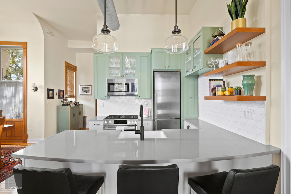 This is an example of a medium sized industrial l-shaped kitchen/diner in Chicago with green cabinets, white splashback, ceramic splashback, stainless steel appliances, light hardwood flooring, a breakfast bar, brown floors and grey worktops.