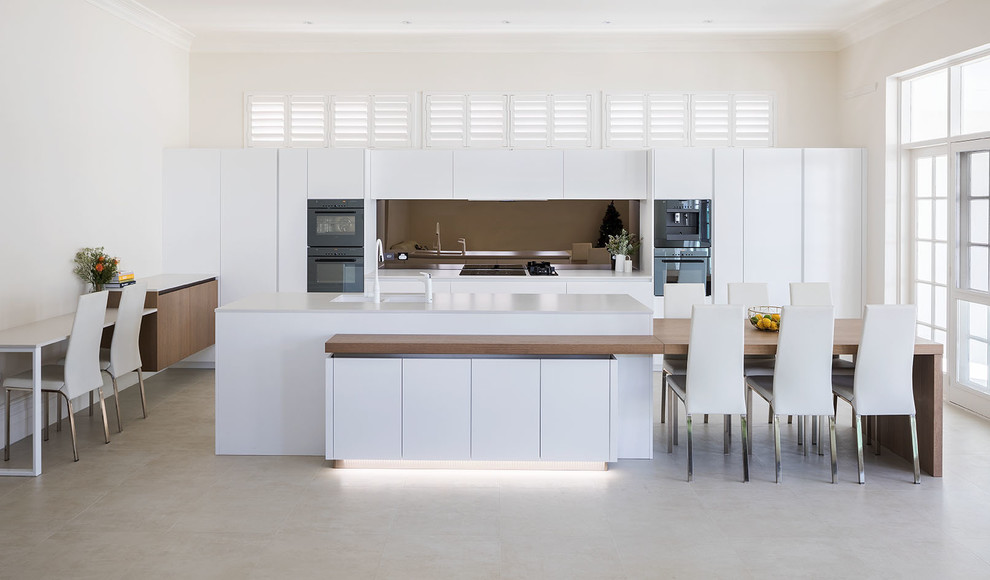 На фото: кухня-гостиная в стиле модернизм с белыми фасадами, столешницей из акрилового камня, фартуком цвета металлик и островом с