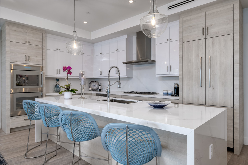 Design ideas for a coastal l-shaped kitchen in Miami with a submerged sink, flat-panel cabinets, white cabinets, white splashback, stone slab splashback, stainless steel appliances, an island, brown floors and white worktops.