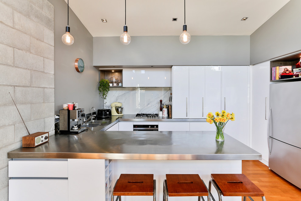 Large industrial u-shaped open plan kitchen in Auckland with a double-bowl sink, flat-panel cabinets, white cabinets, stainless steel worktops, white splashback, glass sheet splashback, stainless steel appliances, medium hardwood flooring, an island and multi-coloured floors.