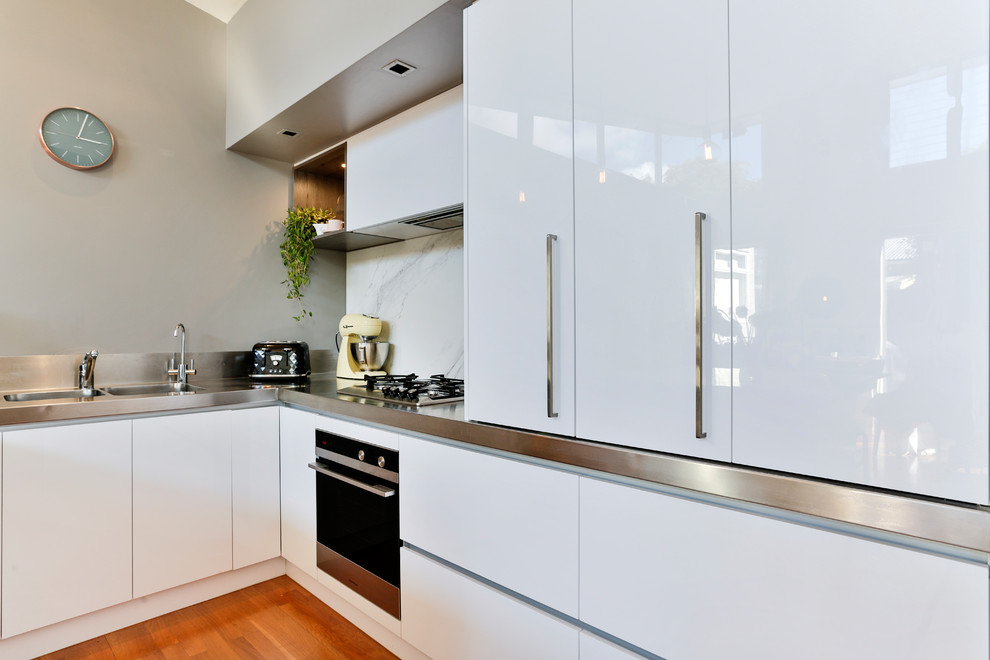 Example of a large urban u-shaped medium tone wood floor and multicolored floor open concept kitchen design in Auckland with a double-bowl sink, flat-panel cabinets, white cabinets, stainless steel countertops, white backsplash, glass sheet backsplash, stainless steel appliances and an island
