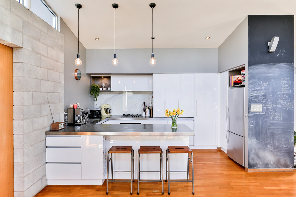 Photo of a large industrial u-shaped open plan kitchen in Auckland with a double-bowl sink, flat-panel cabinets, white cabinets, stainless steel worktops, white splashback, glass sheet splashback, stainless steel appliances, medium hardwood flooring, an island and multi-coloured floors.