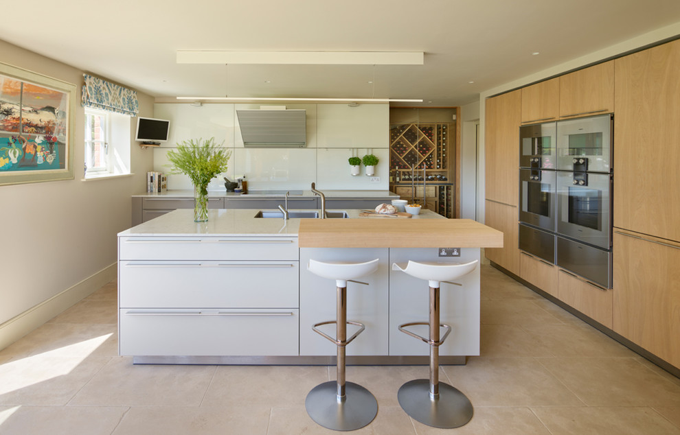 This is an example of a medium sized contemporary l-shaped kitchen/diner in Surrey with a double-bowl sink, flat-panel cabinets, white cabinets, composite countertops, white splashback, glass sheet splashback, stainless steel appliances, an island, beige floors and white worktops.