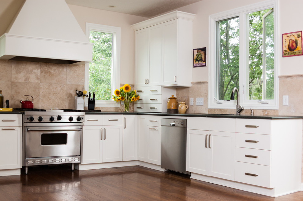 Cette photo montre une cuisine chic en L avec un placard à porte shaker, des portes de placard blanches, une crédence beige, un électroménager en acier inoxydable, un sol en bois brun, un sol marron et plan de travail noir.