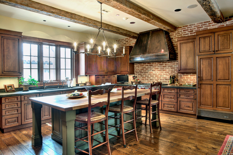 Example of a large tuscan l-shaped dark wood floor and brown floor enclosed kitchen design in Houston with an undermount sink, raised-panel cabinets, dark wood cabinets, soapstone countertops, red backsplash, brick backsplash, paneled appliances, an island and red countertops