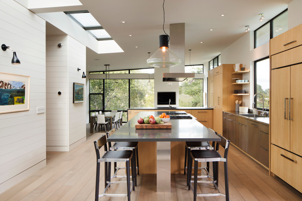 Foto di un cucina con isola centrale design con ante lisce, ante in legno chiaro, elettrodomestici da incasso, top in acciaio inossidabile e parquet chiaro
