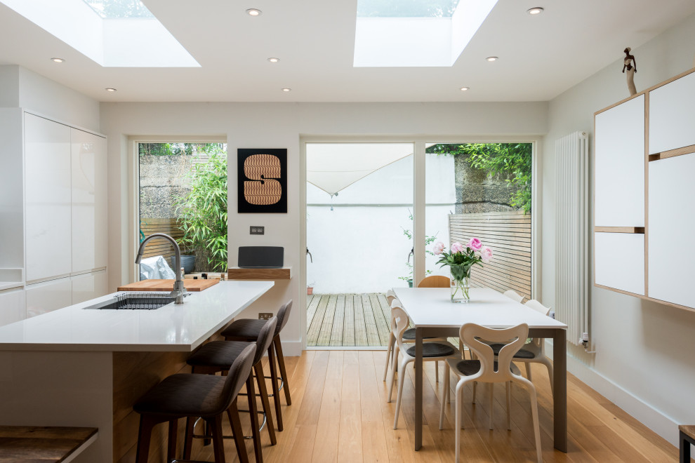 This is an example of a contemporary kitchen/diner in London with a submerged sink, flat-panel cabinets, white cabinets, light hardwood flooring, an island and white worktops.