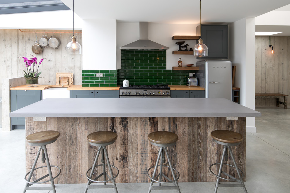 Mid-sized urban single-wall concrete floor and gray floor kitchen photo in Other with a farmhouse sink, recessed-panel cabinets, gray cabinets, green backsplash, subway tile backsplash, stainless steel appliances, an island and wood countertops