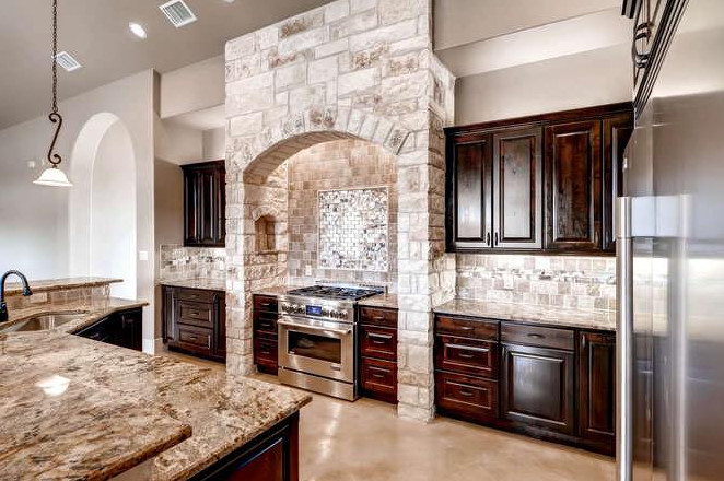 Inspiration for a large mediterranean l-shaped concrete floor eat-in kitchen remodel in Austin with a single-bowl sink, raised-panel cabinets, dark wood cabinets, granite countertops, beige backsplash, stone tile backsplash, stainless steel appliances and an island