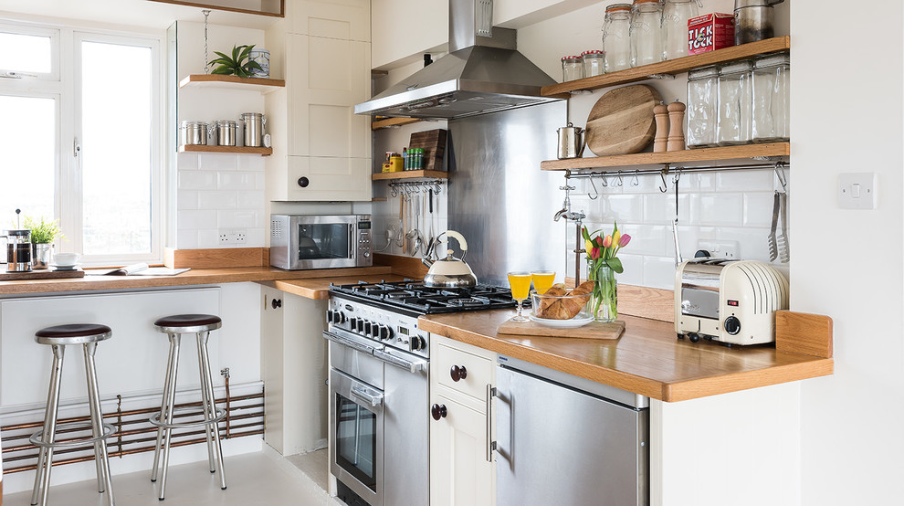 ロンドンにあるインダストリアルスタイルのおしゃれなコの字型キッチン (エプロンフロントシンク、シェーカースタイル扉のキャビネット、ベージュのキャビネット、木材カウンター、白いキッチンパネル、サブウェイタイルのキッチンパネル、シルバーの調理設備) の写真