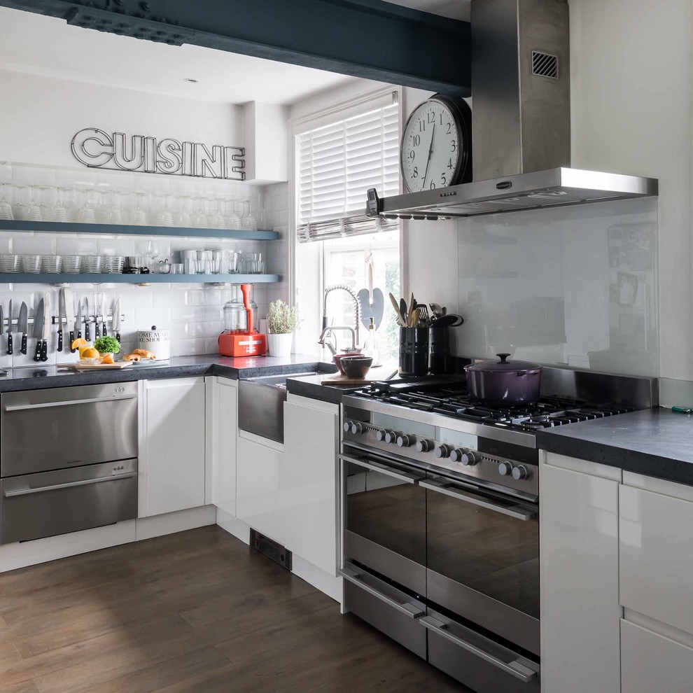 Foto på ett funkis l-kök, med en rustik diskho, släta luckor, vita skåp, grått stänkskydd, glaspanel som stänkskydd, rostfria vitvaror och mörkt trägolv