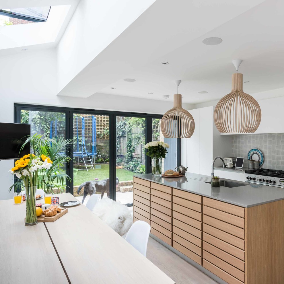 This is an example of a scandi galley kitchen/diner in London with a submerged sink, flat-panel cabinets, white cabinets, grey splashback, stainless steel appliances, medium hardwood flooring and an island.
