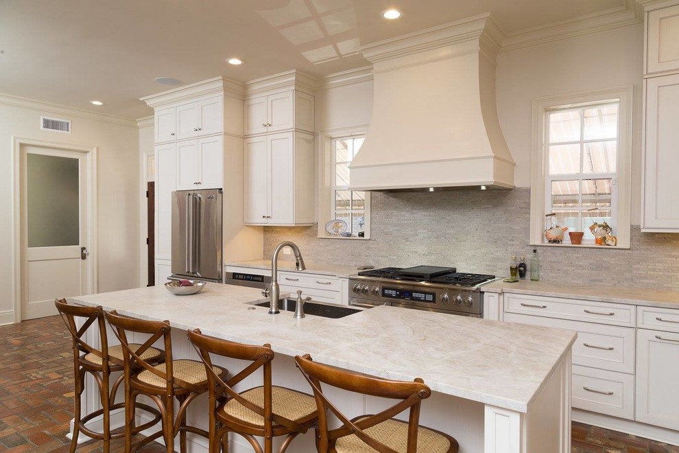 Inspiration for a mid-sized contemporary l-shaped brick floor and multicolored floor kitchen remodel in New Orleans with a farmhouse sink, shaker cabinets, white cabinets, beige backsplash, stone tile backsplash, stainless steel appliances and an island