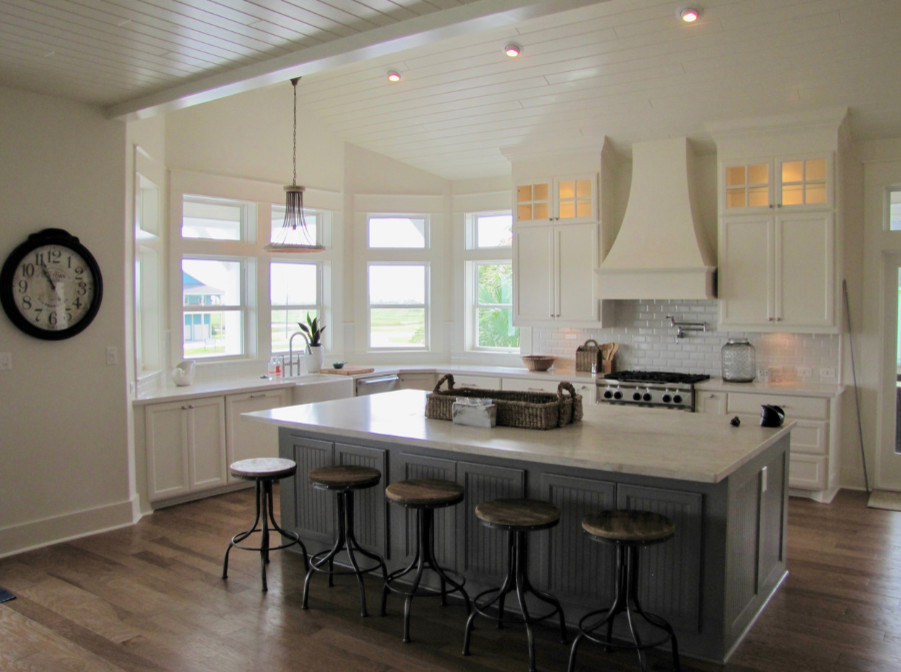 Example of a mid-sized beach style l-shaped medium tone wood floor and brown floor open concept kitchen design in Houston with a farmhouse sink, recessed-panel cabinets, white cabinets, quartz countertops, white backsplash, subway tile backsplash, stainless steel appliances, an island and white countertops