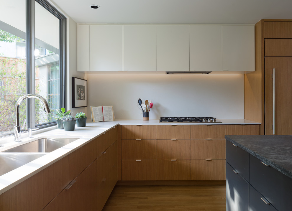 Photo of a midcentury l-shaped kitchen in Austin with a double-bowl sink, flat-panel cabinets, medium wood cabinets, white splashback, integrated appliances, medium hardwood flooring, an island, brown floors and white worktops.