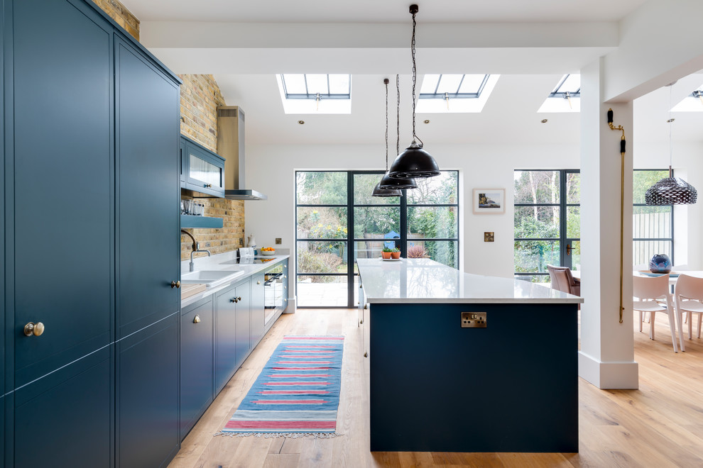 Einzeilige Moderne Wohnküche mit Einbauwaschbecken, flächenbündigen Schrankfronten, blauen Schränken, Rückwand aus Metrofliesen, Küchengeräten aus Edelstahl, braunem Holzboden und Kücheninsel in London