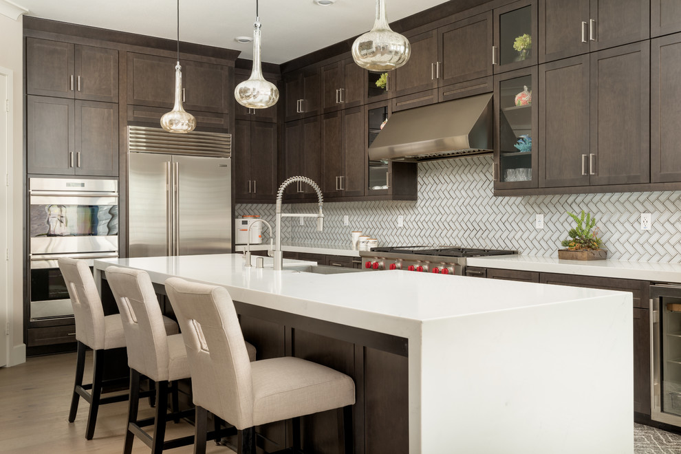 Inspiration for a medium sized classic l-shaped kitchen in Orange County with a belfast sink, shaker cabinets, dark wood cabinets, engineered stone countertops, white splashback, glass tiled splashback, stainless steel appliances, light hardwood flooring, an island, white worktops and beige floors.