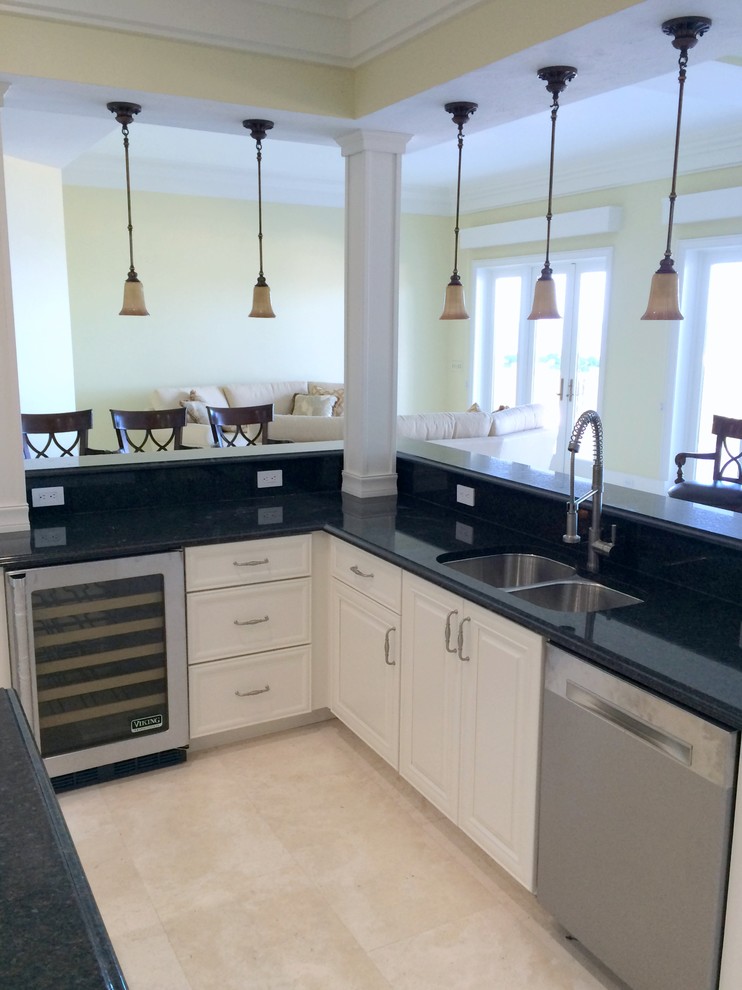 Klassisk inredning av ett stort kök, med en dubbel diskho, luckor med upphöjd panel, vita skåp, granitbänkskiva, svart stänkskydd, stänkskydd i sten, rostfria vitvaror, travertin golv och en köksö
