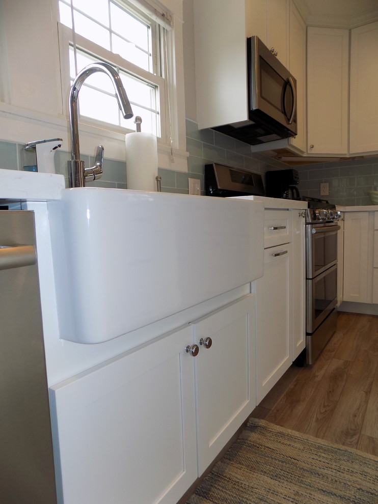 Classic l-shaped kitchen/diner in San Diego with a belfast sink, shaker cabinets, white cabinets, engineered stone countertops, blue splashback, glass tiled splashback, stainless steel appliances, light hardwood flooring, an island and grey floors.