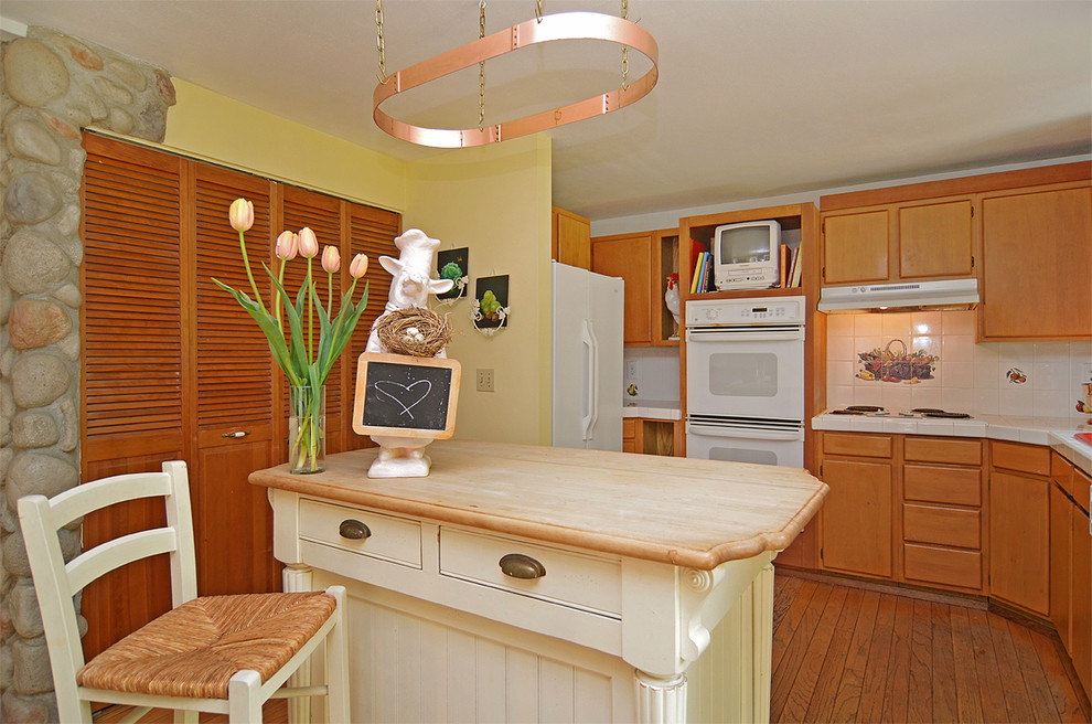 Example of a mid-sized country u-shaped medium tone wood floor open concept kitchen design in Seattle with a drop-in sink, flat-panel cabinets, medium tone wood cabinets, tile countertops, yellow backsplash, ceramic backsplash, white appliances and an island