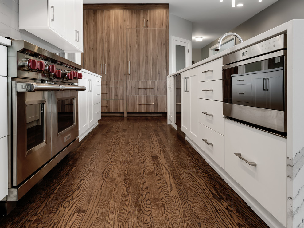 Modern inredning av ett avskilt, stort vit vitt l-kök, med en undermonterad diskho, släta luckor, vita skåp, bänkskiva i kvarts, grått stänkskydd, stänkskydd i porslinskakel, integrerade vitvaror, mellanmörkt trägolv, en köksö och brunt golv