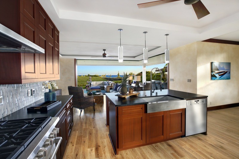 ハワイにある広いトロピカルスタイルのおしゃれなキッチン (エプロンフロントシンク、濃色木目調キャビネット、白いキッチンパネル、ガラスタイルのキッチンパネル、シルバーの調理設備、淡色無垢フローリング) の写真