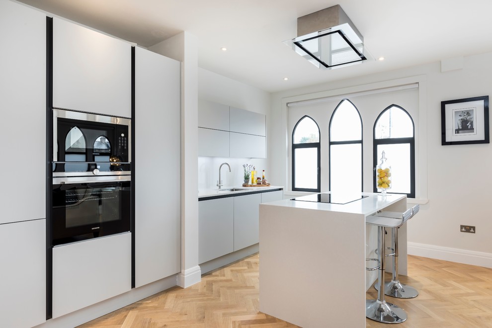 Mid-sized trendy galley light wood floor and beige floor kitchen photo in London with an undermount sink, flat-panel cabinets, an island, white countertops, gray cabinets and black appliances