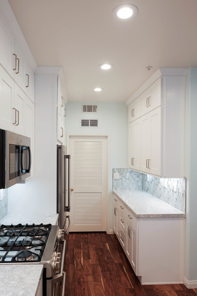 Inredning av ett modernt litet kök, med en nedsänkt diskho, skåp i shakerstil, vita skåp, bänkskiva i kvarts, blått stänkskydd, stänkskydd i glaskakel, rostfria vitvaror, mellanmörkt trägolv och brunt golv