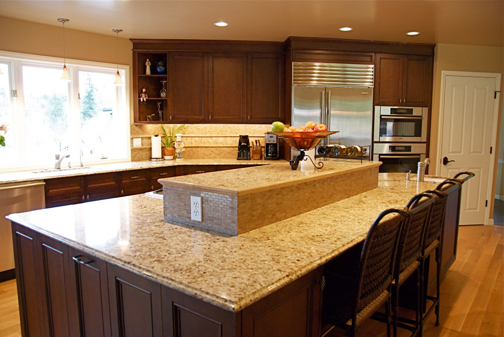 This is an example of an expansive classic l-shaped open plan kitchen in Seattle with a submerged sink, recessed-panel cabinets, dark wood cabinets, granite worktops, beige splashback, travertine splashback, stainless steel appliances, medium hardwood flooring, an island, brown floors and beige worktops.