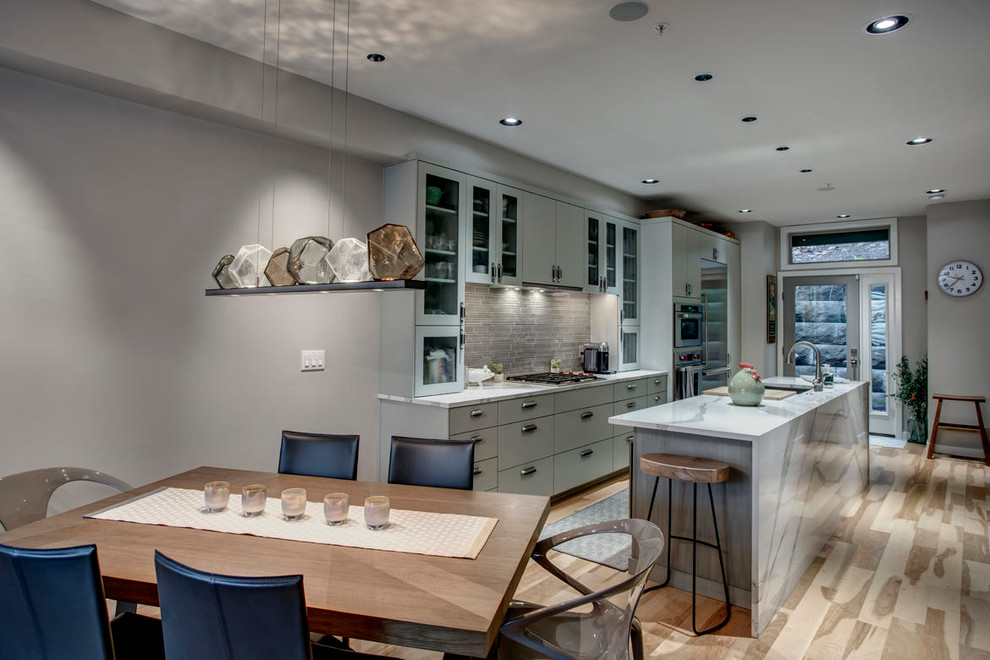 This is an example of a medium sized modern kitchen/diner in Seattle with a submerged sink, glass-front cabinets, grey cabinets, engineered stone countertops, grey splashback, glass tiled splashback, stainless steel appliances, light hardwood flooring, an island, beige floors and white worktops.