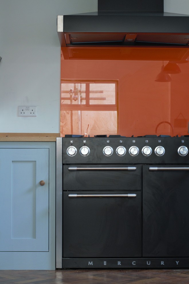 Modelo de cocina de estilo americano de tamaño medio con armarios estilo shaker, puertas de armario turquesas, encimera de madera, salpicadero naranja, salpicadero de vidrio templado, electrodomésticos negros, suelo de madera oscura y una isla