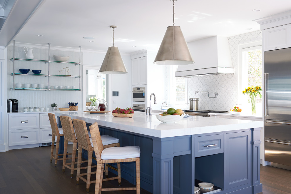 This is an example of a large beach style u-shaped kitchen/diner in New York with a belfast sink, recessed-panel cabinets, blue cabinets, engineered stone countertops, white splashback, ceramic splashback, stainless steel appliances, dark hardwood flooring, an island, brown floors and white worktops.